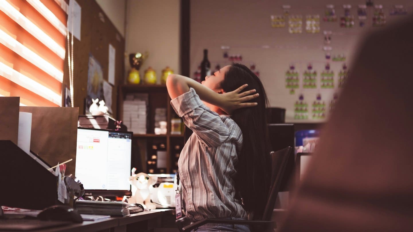 Woman stretches with hands behind her head at an office desk.